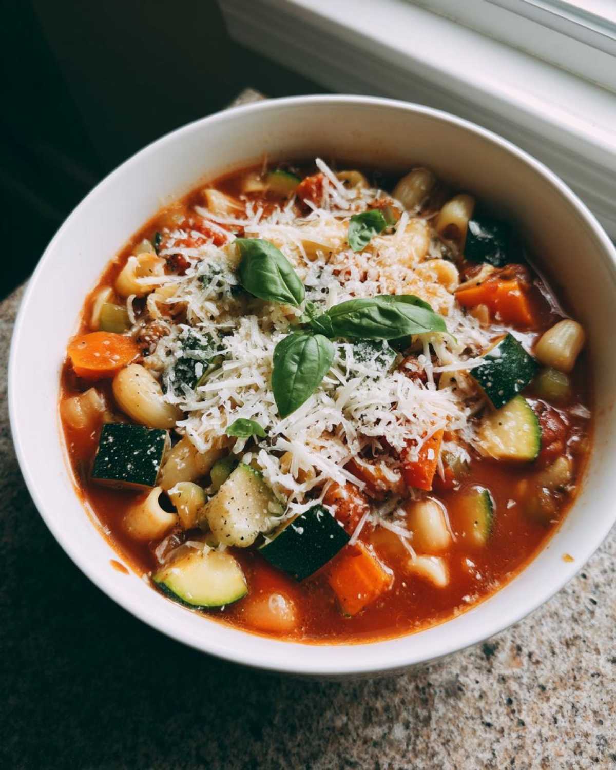 Zbliżenie na miskę Wiosenna zupa minestrone z kawałkami cukinii, marchewki, makaronu i obficie posypana serem.
