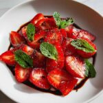 Zbliżenie na talerz z pokrojonymi truskawkami, polanymi sosem balsamicznym i udekorowanymi miętą, czyli Carpaccio z truskawek z octem balsamicznym.