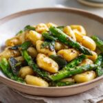 Zbliżenie na miskę pełną Gnocchi ze szparagami w sosie maślanym, ozdobione szałwią.