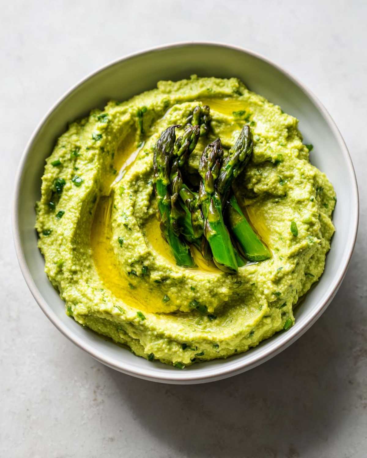 Kremowy, zielony hummus ze szparagami polany oliwą z oliwek w szarej misce.