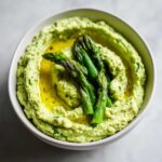 Zbliżenie na miskę kremowego, zielonego Hummus ze szparagami polany oliwą.