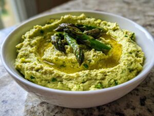 Zielony hummus ze szparagami polany oliwą w białej misce.
