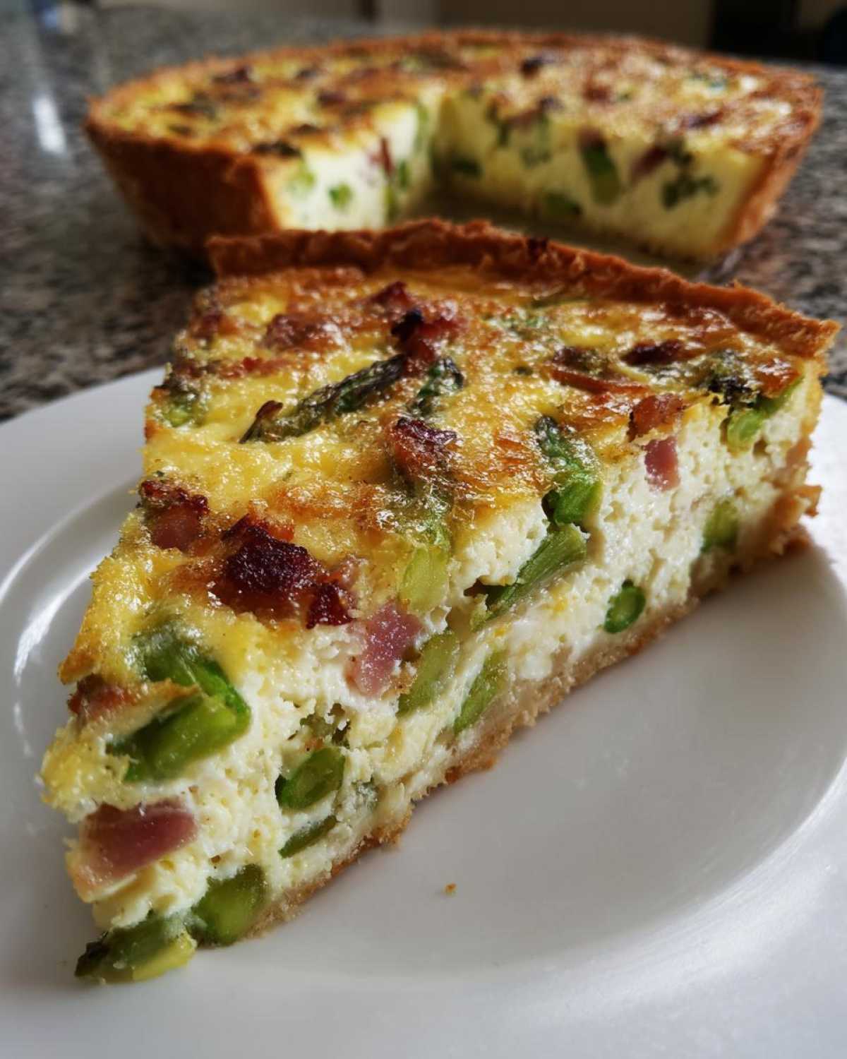 Kawałek pysznego quiche ze szparagami i boczkiem na białym talerzu, w tle widać resztę tarty.