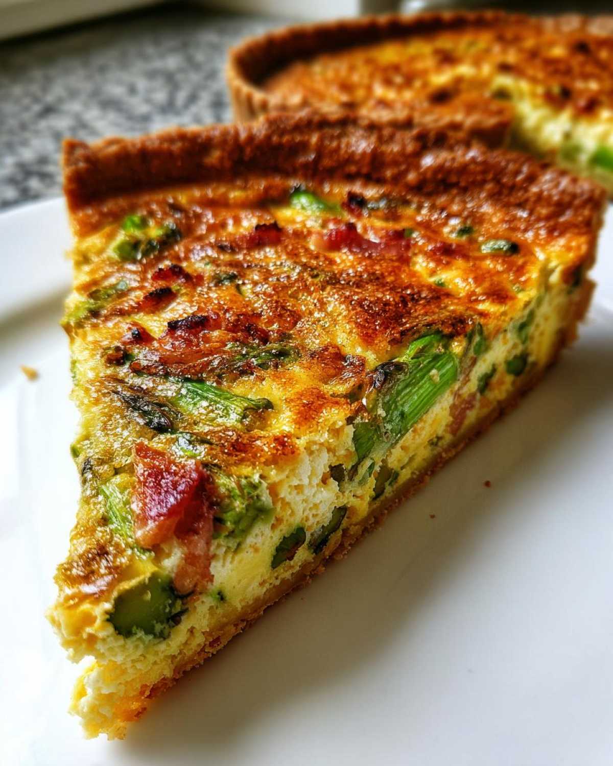 Zbliżenie na kawałek quiche ze szparagami i boczkiem na białym talerzu.