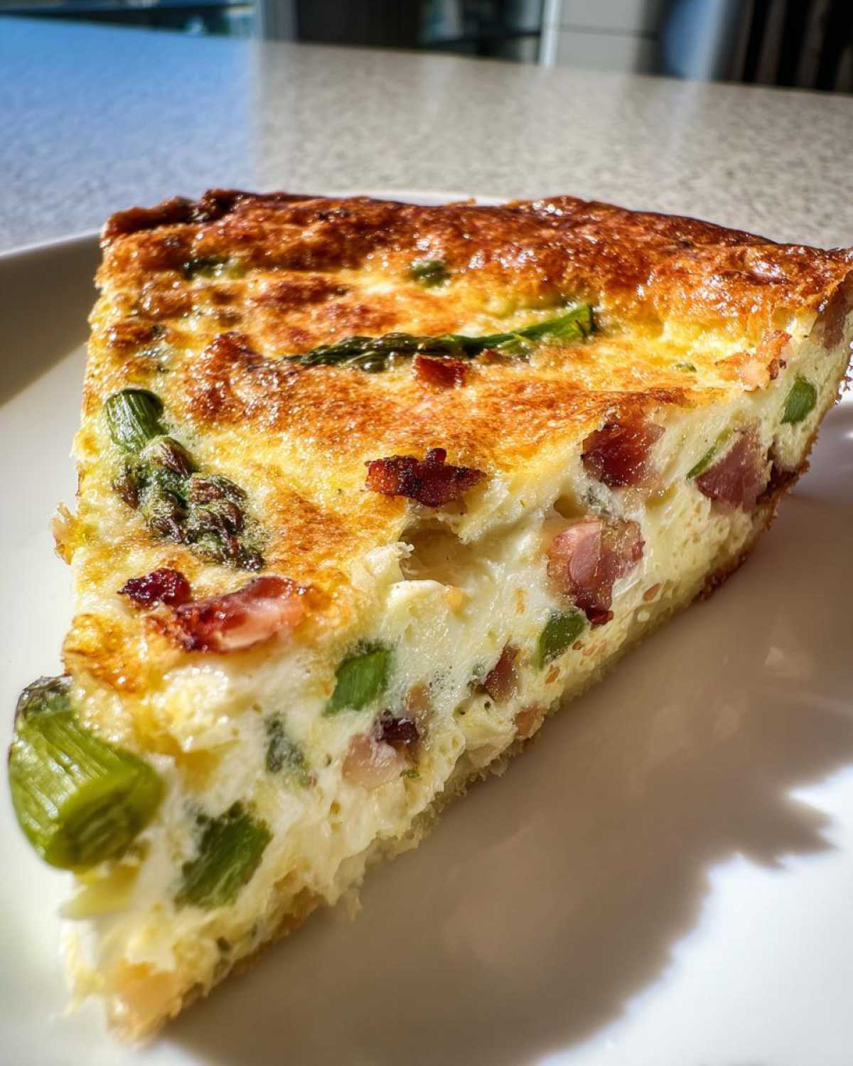 Zbliżenie na kawałek pysznego quiche ze szparagami i boczkiem na białym talerzu.