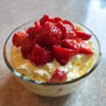 Kremowy kogel-mogel z dużą ilością świeżych, krojonych truskawek na wierzchu w szklanej miseczce.