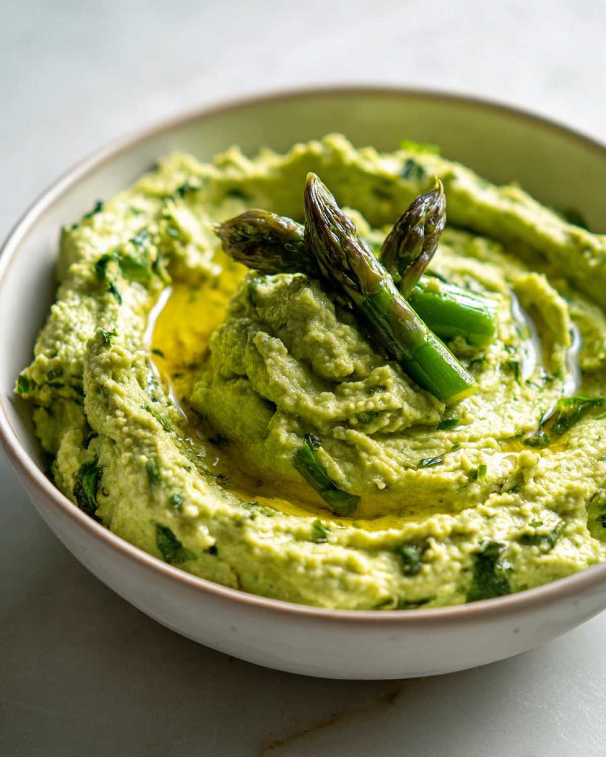 Kremowy, zielony hummus ze szparagami polany oliwą, udekorowany wierzchołkami szparagów.