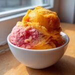 Dwie gałki sorbetu: pomarańczowe lody mango i różowe lody truskawkowe w białej miseczce.
