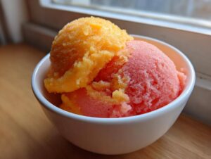 Dwie gałki sorbetu: mango (pomarańczowy) i truskawka (różowy), podane w białej miseczce.
