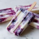 Zbliżenie na mrożone lody na patyku z jogurtu i jagód, ułożone na marmurowym blacie.