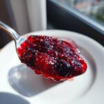 Łyżka pełna gęstego, błyszczącego dżemu truskawkowego z borówką i chia, na białym talerzyku.