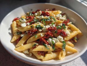 Miseczka makaronu penne z bobem, suszonymi pomidorami i serem feta, posypana bazylią.