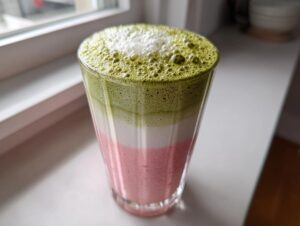 Warstwowe Matcha latte z musem truskawkowym, z zieloną pianką matcha na wierzchu, stojące na parapecie.