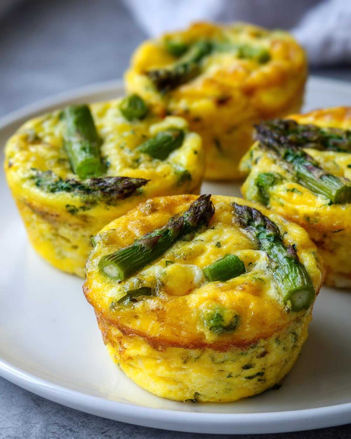 Zbliżenie na cztery puszyste muffinki jajeczne ze szparagami i serem na białym talerzu.