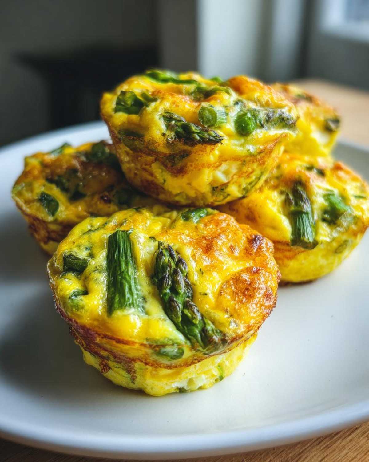Zbliżenie na stos puszystych muffinek jajecznych ze szparagami i serem na białym talerzu.