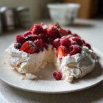 Przekrojona Pavlova z owocami jagodowymi, bitą śmietaną i cukrem pudrem na białym talerzu.