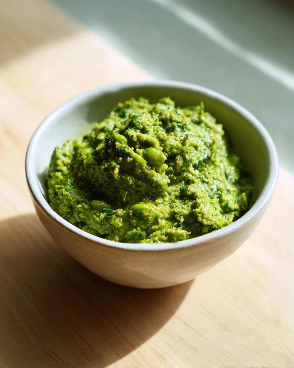 Gęste, zielone Pesto z bobu z miętą i cytryną w małej, jasnej miseczce na drewnianym blacie.