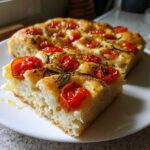 Kawałek puszystej Foccacia z pomidorkami i rozmarynem na białym talerzu, widoczna puszysta struktura ciasta.