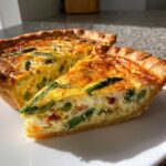 Kawałek pysznego Quiche ze szparagami i boczkiem na białym talerzu, z widocznym złocistym spodem.