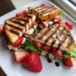 Trójkątne kawałki grillowanego sera halloumi z truskawkami i sałatą, polane sosem balsamicznym, jako Sałatka z grillowanym halloumi i truskawkami.