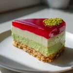 Kawałek sernika pistacjowego na zimno z malinami, z warstwą galaretki i posypką z pistacji.