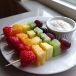 Trzy tęczowe szaszłyki owocowe z truskawkami, melonem, ananasem, kiwi i winogronami obok miseczki jogurtu.