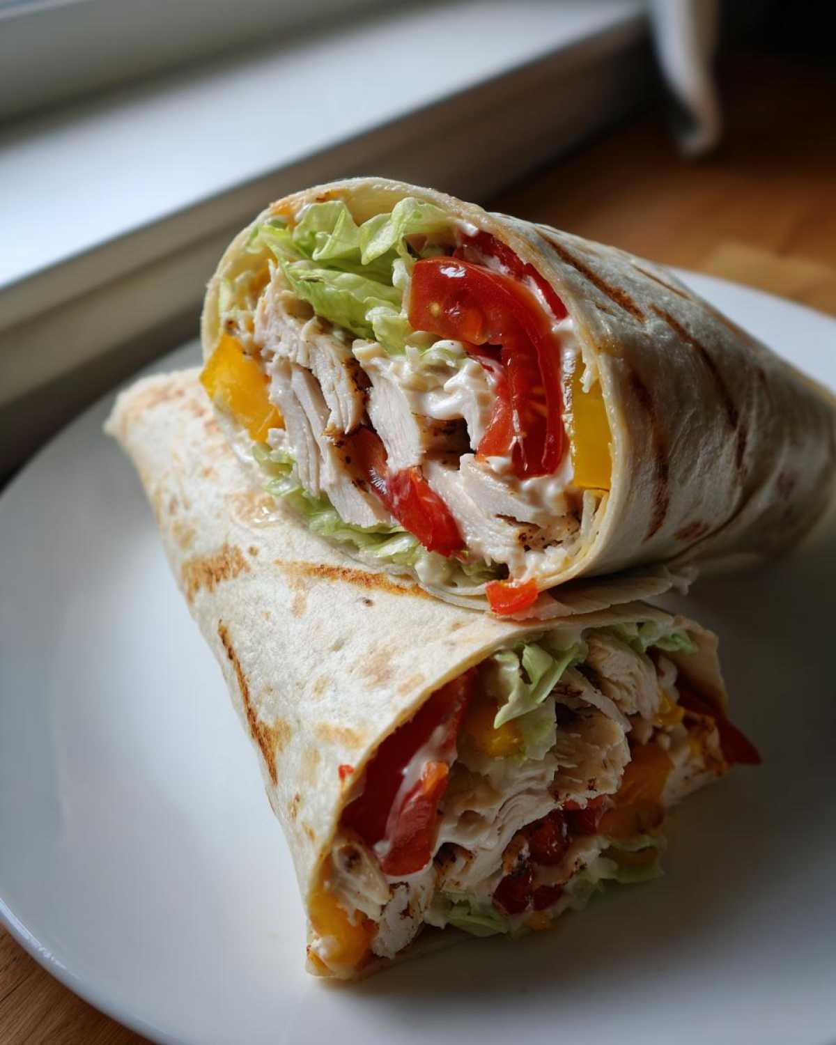 Przekrojona na pół tortilla z grillowanym kurczakiem, sałatą, pomidorem i żółtą papryką na białym talerzu.